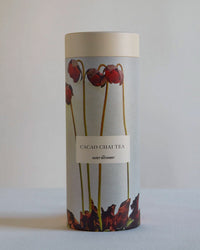 The Quiet Botanist Cacao Chai Tea Gift Set comes in a cylindrical container with an illustrated design of tall red flowers and leaves on a light gray background.