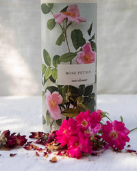 A cylindrical container of The Quiet Botanist Edible Rose Petals, decorated with botanical rose illustrations, stands behind a small pile of vibrant organic petals—ideal for cake decoration—on a white surface.