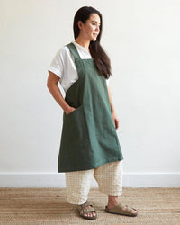 A woman stands sideways on a woven rug against a white wall, wearing The Floral Society's Canvas Workshop Apron in Fern over a white shirt, loose checkered pants, and tan sandals. The pure cotton apron features large pockets and a cross-back design.