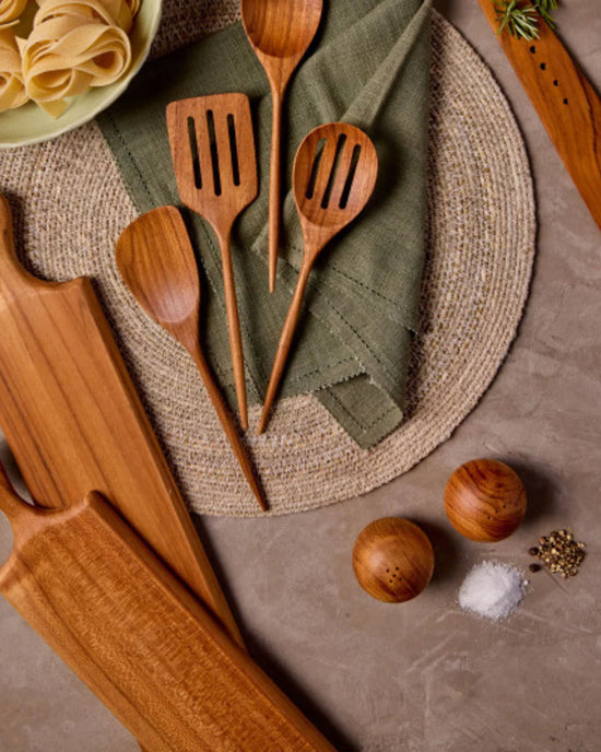 Be Home US Teak Salt & Pepper Shaker Set, wooden kitchen utensils, cutting boards, and a bowl of pasta are displayed on a woven placemat atop a gray countertop.