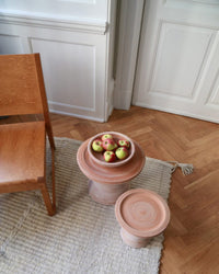A bowl of apples rests on a round wooden side table near a Bergs Potter Celeste 9 inch Pedestal Planter in Raw Rosa, a wooden chair, and a textured rug atop a herringbone wood floor.