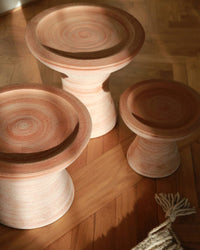Three Celeste 9 inch Pedestal Planters in Raw Rosa by Bergs Potter, featuring a handcrafted pottery-inspired concentric circular design, are arranged on a wooden floor beside a fringed textile.