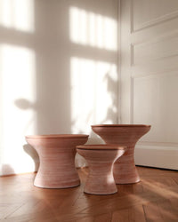 Three wooden, hourglass-shaped stools of varying heights sit on a parquet floor near a white wall and door, beside the Bergs Potter Celeste 9 inch Pedestal Planter in Raw Rosa.
