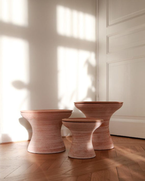 Three wooden, hourglass-shaped stools of varying heights sit on a parquet floor near a white wall and door, beside the Bergs Potter Celeste 9 inch Pedestal Planter in Raw Rosa.