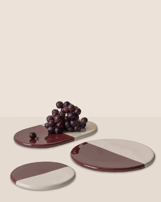 Three Bergs Potter Nuna Glazed 11 inch Trays in Sandstone/Deep Burgundy are arranged on a beige surface, with a cluster of red grapes placed on the largest tray.