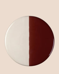 The Bergs Potter Nuna Glazed 11 inch Tray features a round stoneware design divided vertically into equal halves: one in sandstone, the other in deep burgundy, set against a light beige background.