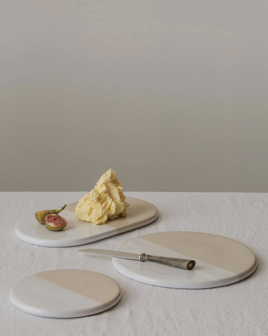 A Nuna Glazed 13.4x7.9 Tray in Sandstone/Mineral White by Bergs Potter, holding a knife, fig, and butter, is displayed with Bergs Potter glazed stoneware coasters on a white tablecloth against a neutral background.