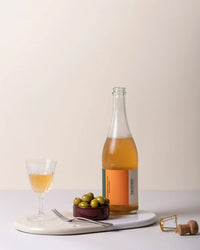 A glass and bottle of orange-hued drink sit on glazed stoneware coasters with a dish of green olives, fork, and cork on the Bergs Potter Nuna Glazed 13.4x7.9 Tray in Sandstone/Mineral White against a neutral background.