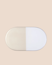 The Bergs Potter Nuna Glazed 13.4 x 7.9 Tray in Sandstone/Mineral White features an oval ceramic design split evenly into beige and white, set against a light beige background.