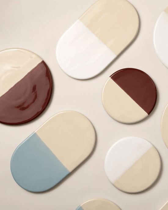 The Bergs Potter Nuna Glazed 7.9 inch Tray in Sandstone/Deep Burgundy features a two-tone round stoneware design, food safe for easy dining, and is shown arranged on a neutral background with other assorted plates.
