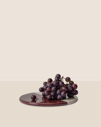 A bunch of dark purple grapes sits on the Bergs Potter Nuna Glazed 7.9 inch Tray in Sandstone/Deep Burgundy, set against a plain beige background.