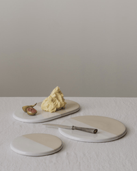 Three round marble serving boards with a butter slab, half a fig, and a silver knife sit on a white textured tablecloth with the Bergs Potter Nuna Glazed 7.9 inch Tray in Sandstone/Mineral White against a plain background.