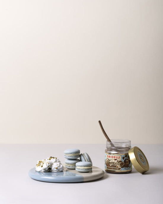 A Bergs Potter Nuna Glazed 7.9 inch Tray in Sandstone/Misty Blue holds blue and white macarons and meringues beside an open hazelnut spread jar with a spoon, all elegantly arranged on glazed stoneware against a plain background.