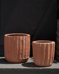 Two empty, fluted terracotta planters by Bloomist—a medium and a small—sit side by side on a dark stone surface against a black wall. These handmade Fluted Planters are perfect for indoor or outdoor use.