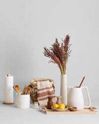 Featuring the Bloomist Hammer Grinder in teak, this curated kitchen set—including a paper towel holder, wooden utensils, basket, towel, vase with dried flowers, pitcher, lemons, and small jars—highlights Danish design against a plain background.