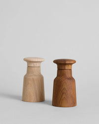 Two Hammer Grinders by Bloomist, in teak and light wood finishes, stand upright on a plain surface. Their smooth cylindrical shapes and rounded tops evoke classic Danish salt and pepper grinder design.