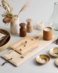 A tabletop arrangement with wooden boards, utensils, brass dishes, a candle, a vase of dried flowers, and the Bloomist Hammer Grinder in teak showcases Danish design against a neutral background.