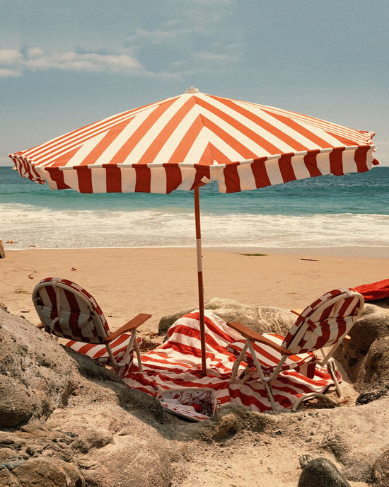 Two striped beach chairs and a Business & Pleasure Co Amalfi Umbrella - Crew Le Sirenuse Stripe stand on the sand facing the ocean, with rocks in front and waves rolling in beneath a partly cloudy sky.