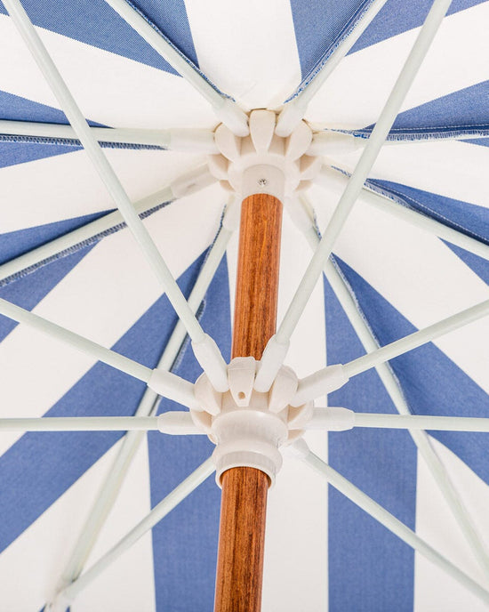 View from beneath the Amalfi Umbrella - Crew Mediterranean Blue Stripe by Business & Pleasure Co, showcasing its blue and white stripes, sturdy wooden pole, and white support spokes for generous sun shade.