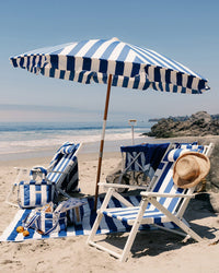 Two blue-and-white striped beach chairs and the Amalfi Umbrella - Crew Mediterranean Blue Stripe by Business & Pleasure Co are set up on sandy beach near the ocean, with a beach bag, cooler, fruit, straw hat, rocks, and calm waves nearby.