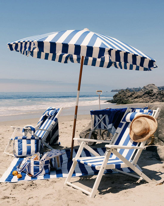 Two blue-and-white striped beach chairs and the Amalfi Umbrella - Crew Mediterranean Blue Stripe by Business & Pleasure Co are set up on sandy beach near the ocean, with a beach bag, cooler, fruit, straw hat, rocks, and calm waves nearby.