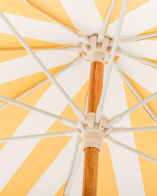 Close-up of the Amalfi Umbrella - Crew Mimosa Yellow Stripe by Business & Pleasure Co, showing its yellow and white UV-resistant canopy, wooden pole, and white spokes radiating outward.