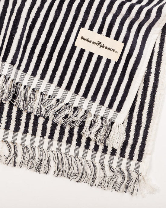 The Beach Blanket - Laurens Navy Stripe by Business & Pleasure Co is a family-friendly, black and white striped, 100% cotton blanket with fringed edges and a sewn-on brand label.