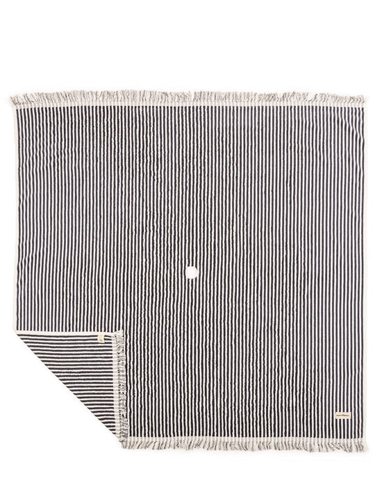 The Beach Blanket - Laurens Navy Stripe by Business & Pleasure Co is a family-friendly, square black and white striped 100% cotton blanket with fringed edges, a small white circle in the center, and a corner folded to reveal the reverse side.