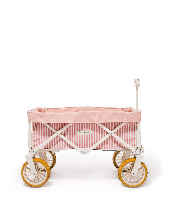 The Business & Pleasure Co Beach Cart - Laurens Pink Stripe features pink and white stripes, yellow sand wheels, a white handle, and a compact foldable design. Photographed against a white background.