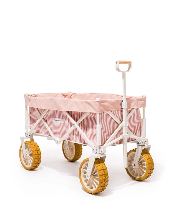 The Beach Cart - Laurens Pink Stripe by Business & Pleasure Co features a compact, foldable design with pink and white striped fabric, a white frame, and large yellow sand wheels, shown against a white background.