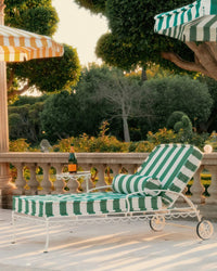 A Bolster Pillow - Crew Canyon Green Stripe by Business & Pleasure Co sits on a sunny patio lounge chair beside a small table with a bottle and glass, surrounded by lush trees and a stone balustrade.