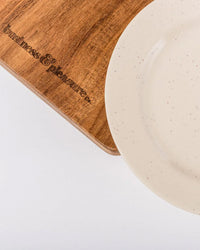 A partial view of the white-speckled plate from the Picnic Set - Laurens Pink Stripe by Business & Pleasure Co sits on a wooden board with engraved text, both arranged on a white surface.
