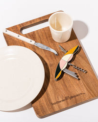 The Picnic Set - Laurens Pink Stripe by Business & Pleasure Co includes a wooden board with a white plate, white-handled cheese knife, colorful corkscrew, and beige cup—ideal for any stylish outdoor picnic.