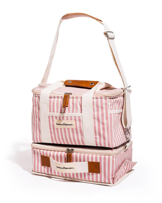 The Picnic Set - Laurens Pink Stripe by Business & Pleasure Co is a pink and white striped soft cooler bag with white handles, brown accents, an adjustable shoulder strap, and two vertical zippered compartments—ideal for picnics.