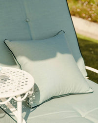 A Small Square Throw Pillow in Rivie Green by Business & Pleasure Co rests on a matching lounge chair next to a white metal outdoor table in a sunlit garden setting.