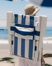 A person in a straw hat carries the Business & Pleasure Co Tommy Chair - Acacia White - Crew Mediterranean Blue Stripe folded on their back while standing on a sandy beach near the ocean.