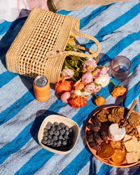 The Canasta Chica Bag in Natural by caminito, a bouquet of flowers, a canned drink, blueberries in a bowl, and a plate of crackers, dried fruits, and cheese rest on a blue-striped blanket for a picnic.