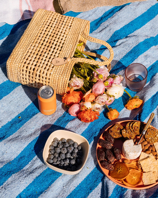 The Canasta Chica Bag in Natural by caminito, a bouquet of flowers, a canned drink, blueberries in a bowl, and a plate of crackers, dried fruits, and cheese rest on a blue-striped blanket for a picnic.