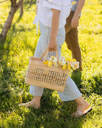 Wearing striped pants and sandals, a person walks barefoot on grass, carrying the caminito Canasta Chica Bag in Natural filled with yellow and white flowers. Another person is nearby, mostly out of frame.