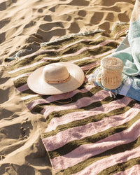 A straw hat and woven basket rest on the caminito Marea Fresca - Blanket Roll, a striped pink and green indoor-outdoor blanket spread over sunlit sand, with part of a light blue towel nearby.