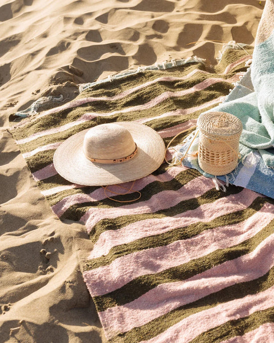 A straw hat and woven basket rest on the caminito Marea Fresca - Blanket Roll, a striped pink and green indoor-outdoor blanket spread over sunlit sand, with part of a light blue towel nearby.