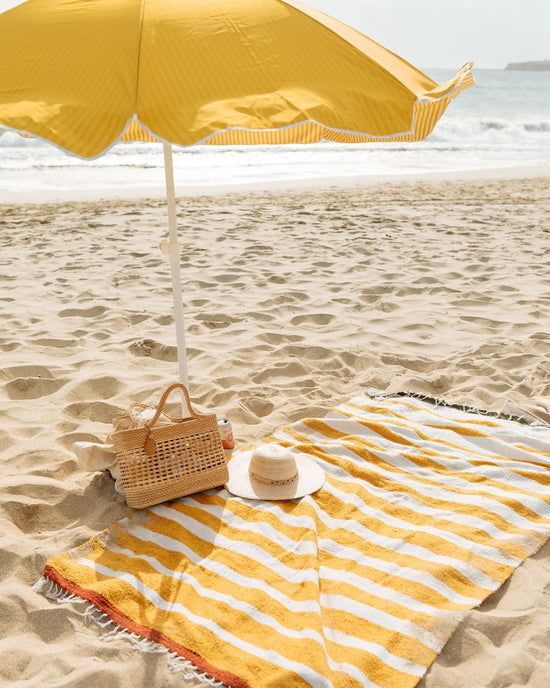 The caminito Marea Sol - Blanket Roll in yellow-and-white stripes is spread on the sandy beach, shaded by a matching umbrella. A woven bag and straw hat sit atop the blanket, with the ocean visible in the background.