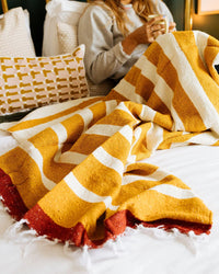 A person sits on a bed with the caminito Marea Sol - Blanket Roll, its yellow, white, and red stripes adding warmth. They hold a cup, and decorative pillows in the background complete this cozy, inviting scene.