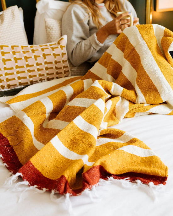 A person sits on a bed with the caminito Marea Sol - Blanket Roll, its yellow, white, and red stripes adding warmth. They hold a cup, and decorative pillows in the background complete this cozy, inviting scene.