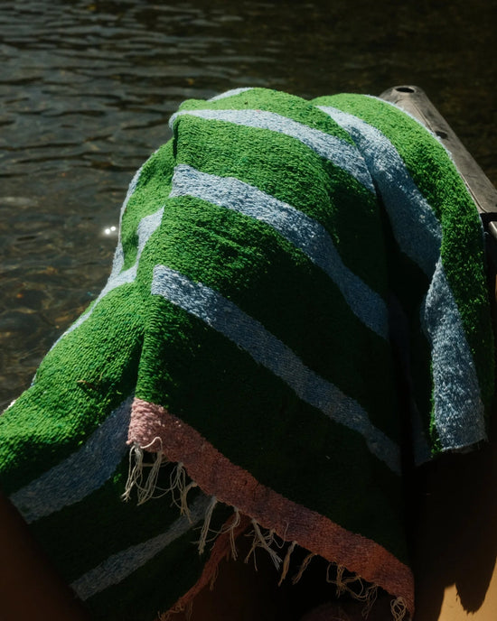 The Marea Vuelta - Blanket Roll by caminito, handwoven in Mexico with green, light blue, and pink stripes and frayed edges, is draped over a surface by water in bright sunlight.