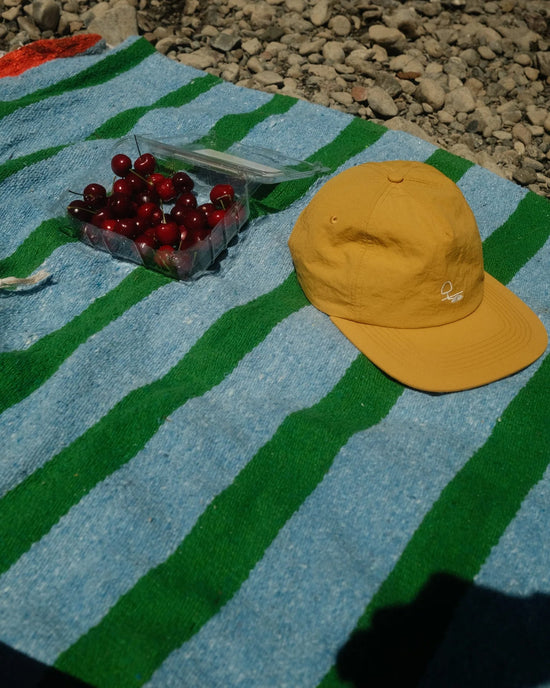 A yellow cap and a container of cherries rest on a caminito Marea Vuelta - Blanket Roll, featuring blue and green stripes, spread over rocks in the bright sunlight.