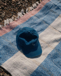 A blue baseball cap rests on a caminito Suave Aire - Blanket Roll—an indoor-outdoor blanket with blue, white, and pink stripes—spread out on a pebble-covered surface.
