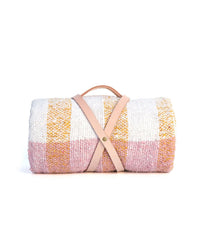 The caminito Suave Miel - Blanket Roll, a handwoven checkered throw in pink, white, and yellow from Mexico, is rolled up and fastened with a handmade leather strap against a white background.