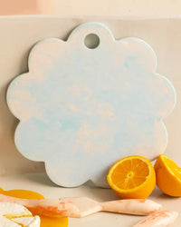 The Casa Amarosa 9in Scallop Resin Cheese/Charcuterie Board in Blue, featuring a flower-like shape and top hole, stands upright behind lemon halves and utensils, bringing charm to your handmade cheese board collection.