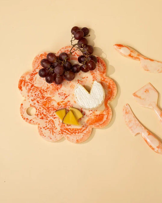 A Casa Amarosa 9in Scallop Resin Cheese/Charcuterie Board in Ivory Pink displays red grapes, a wheel of white cheese, and two yellow cheese slices. Matching marbled cheese utensils are arranged on the right.
